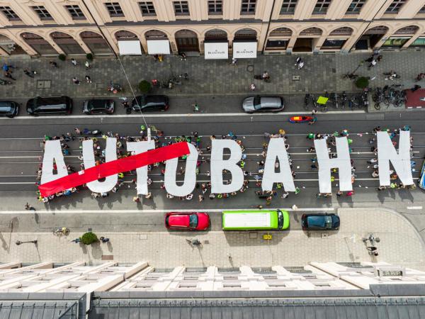 Foto von oben auf Menschen, die Buchstaben hochhalten: AUTOBAHN - das Wort Auto ist durchgestrichen