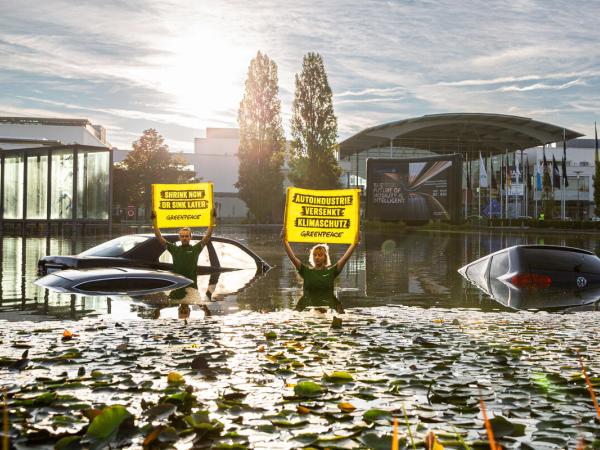Protest vor IAA. Autodächer im Wasser, 2 Aktive halten Banner: "Shrink now or swim later",  "Autoindustrie versenkt Klimaschutz"