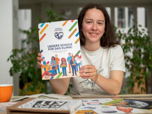 Eine junge Frau sitzt am Schreibtisch und hält die Handreichung "Unsere Schule für das Klima" in die Kamera